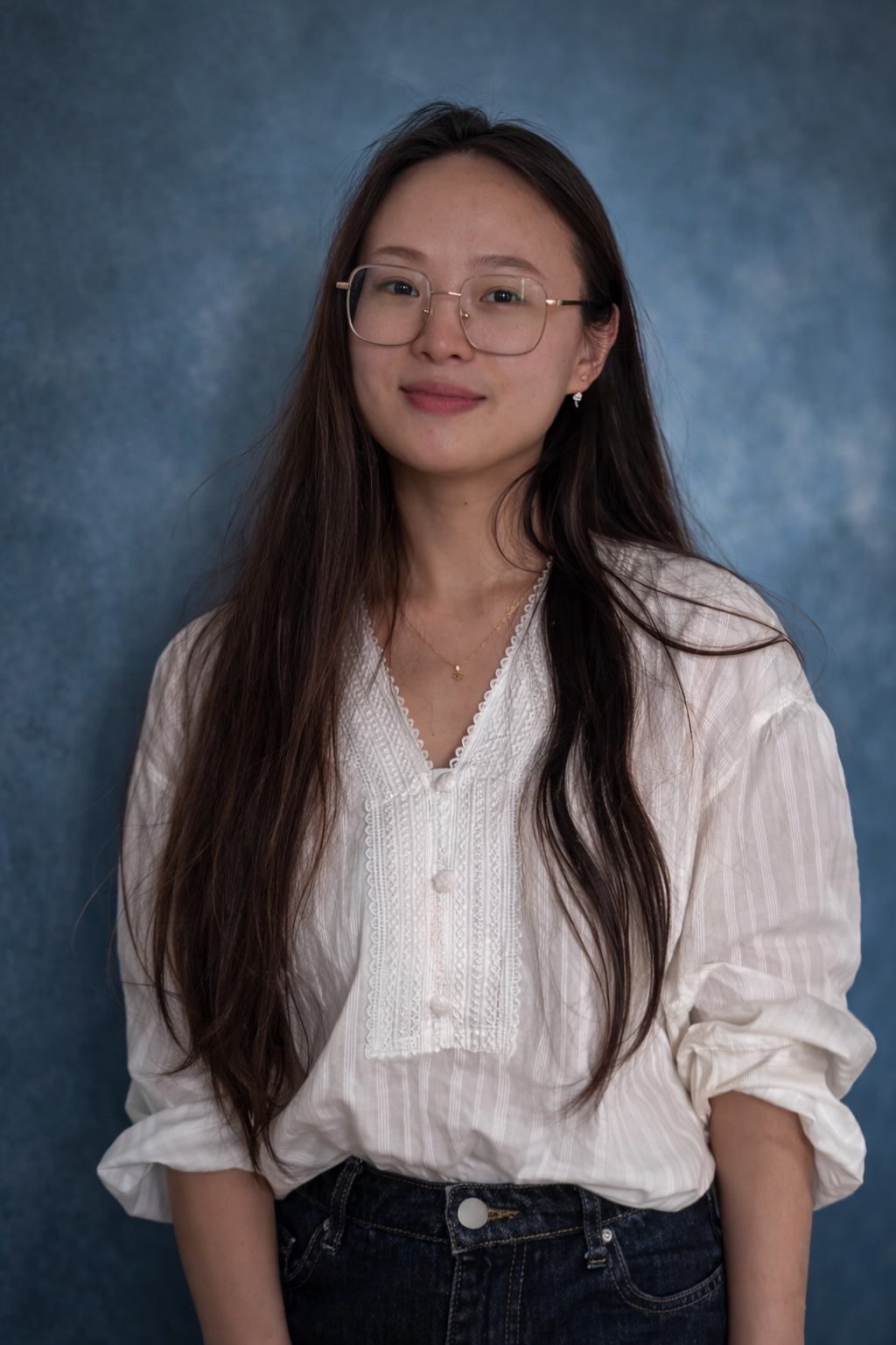 A smiling young woman with long dark hair, wearing a white button-up shirt with a lace detail, standing in front of a soft blue background.