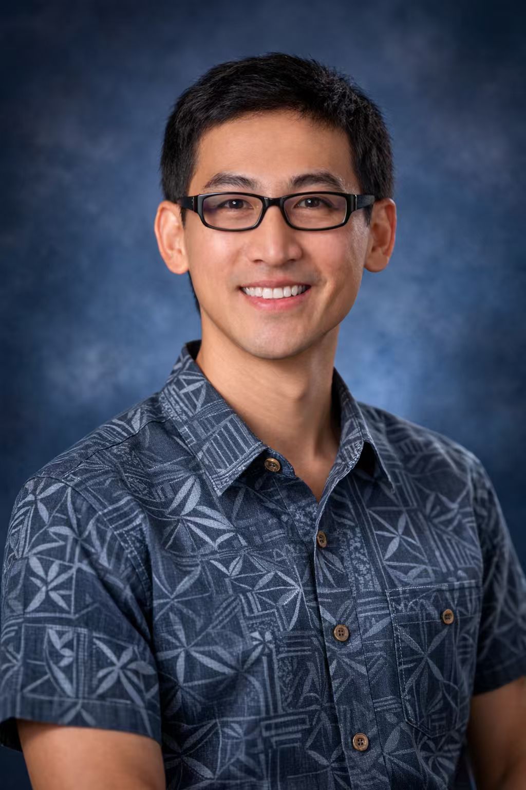 Portrait of a man wearing glasses and a patterned short-sleeve shirt, smiling against a soft blue background.